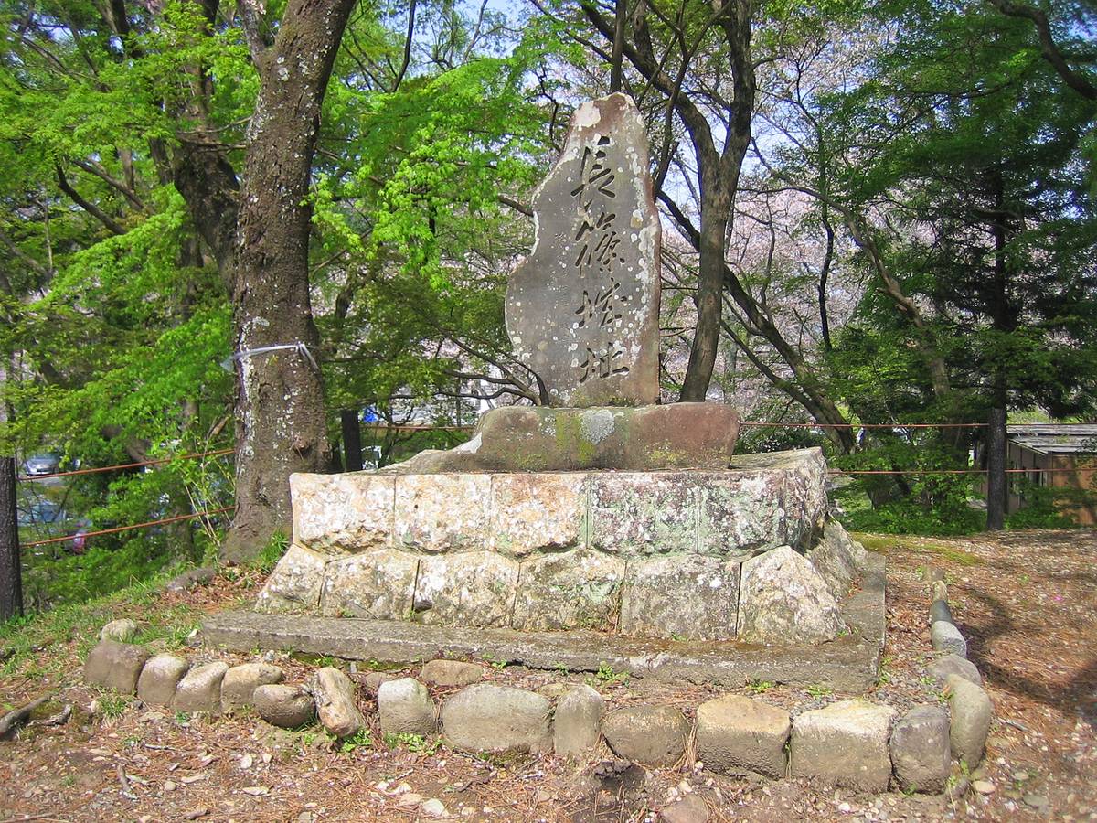 A stone monument with Japanese characters on a stone base, surrounded by trees with green leaves and pink cherry blossoms.