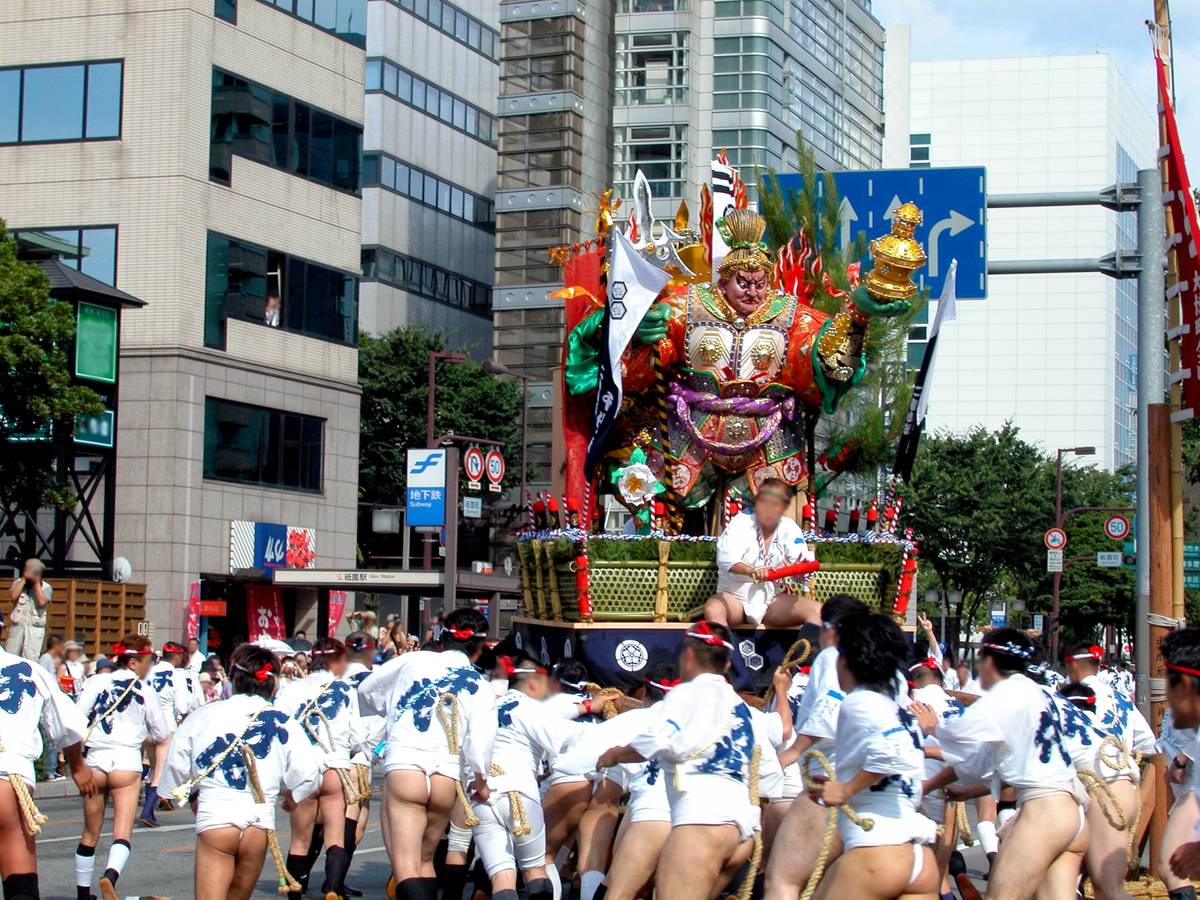 Men in traditional loincloths pull a large, colorful float with a warrior deity through a city street during a Japanese festival.