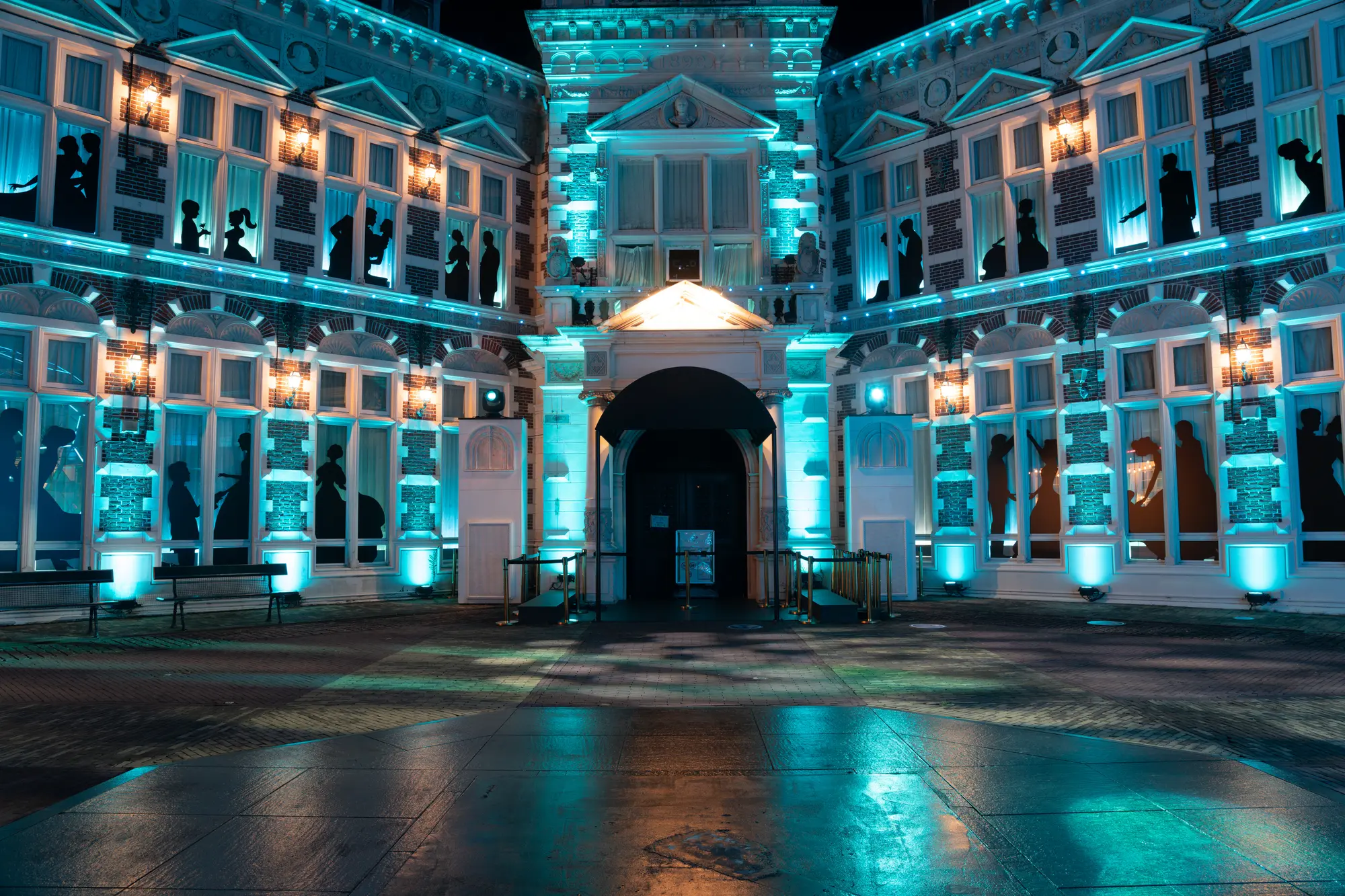 Night view of a building illuminated with teal and warm lights, featuring silhouettes of people in its windows.