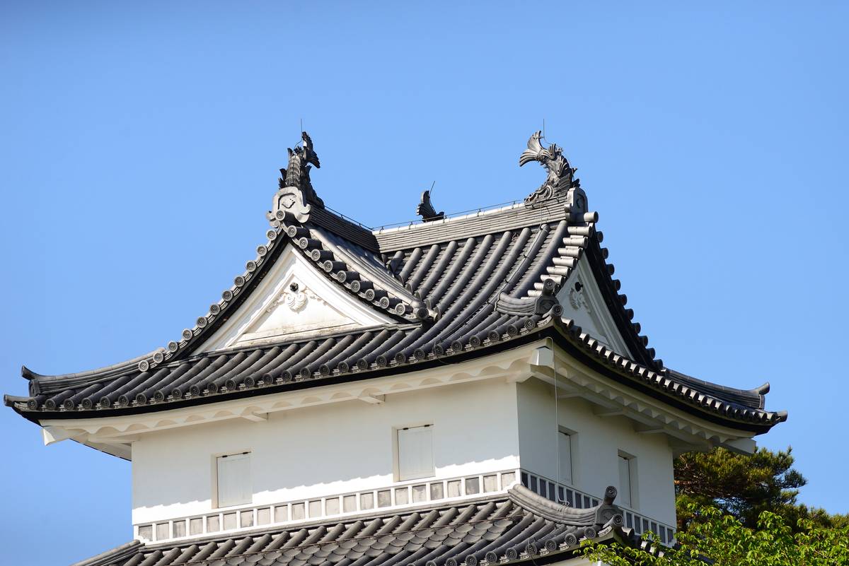 White Japanese castle tower with a multi-layered dark tiled roof, featuring decorative finials, against a clear blue sky.