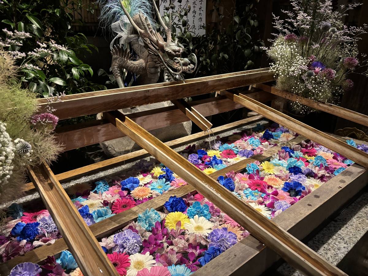 A vibrant Japanese purification fountain with colorful floating flowers, wooden water channels, and a dragon statue.