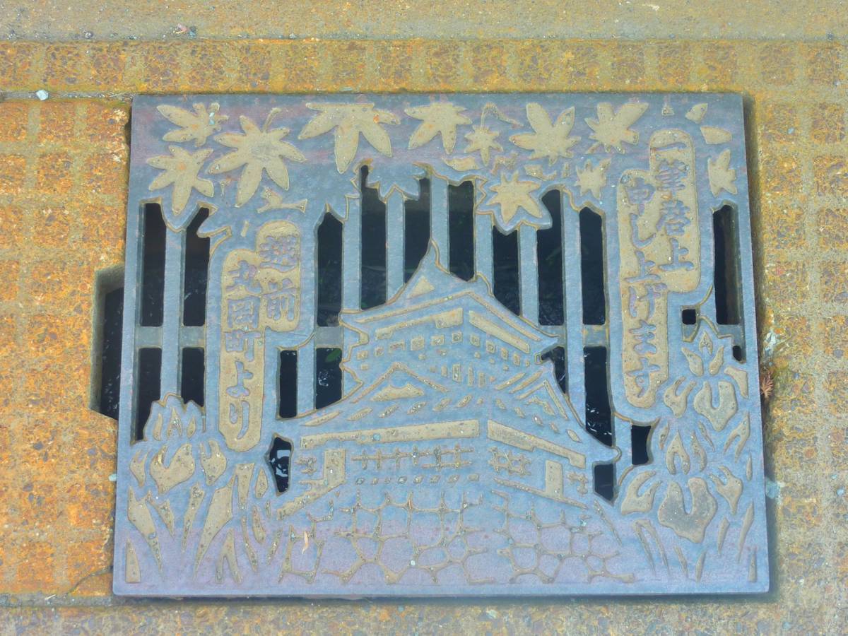 An ornate metal grate depicting a Japanese castle, maple leaves, and vertical Japanese script, embedded in rusty paving.
