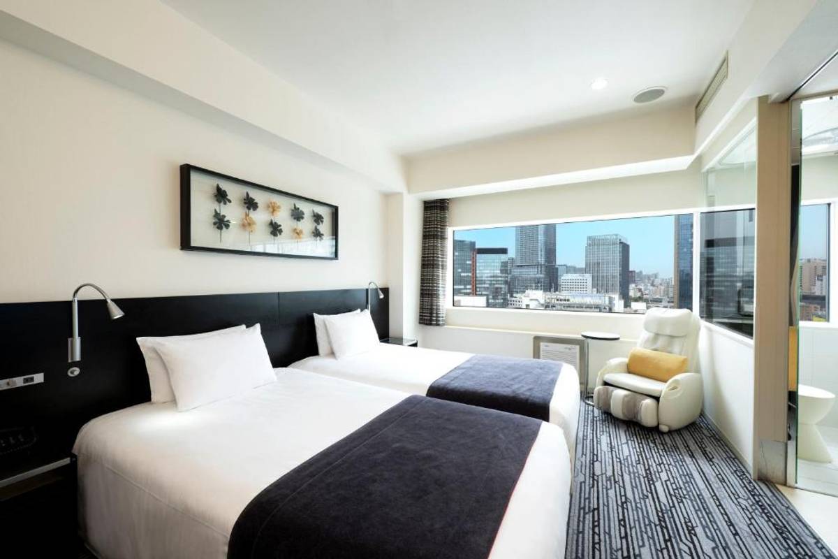 A hotel room with two twin beds, a city view from a large window, and a white massage chair.