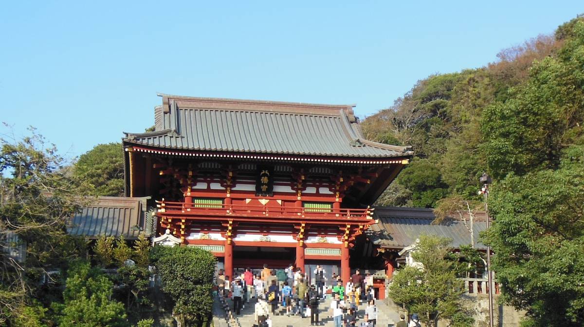 A red Japanese temple gate with a grey roof, crowded with people, surrounded by lush green trees under a clear blue sky.