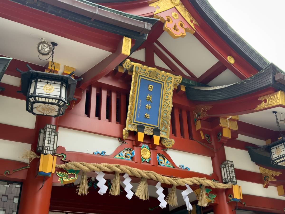 The entrance of Hie Shrine, featuring red and gold traditional Japanese architecture, lanterns, and a braided rope with white paper streamers.