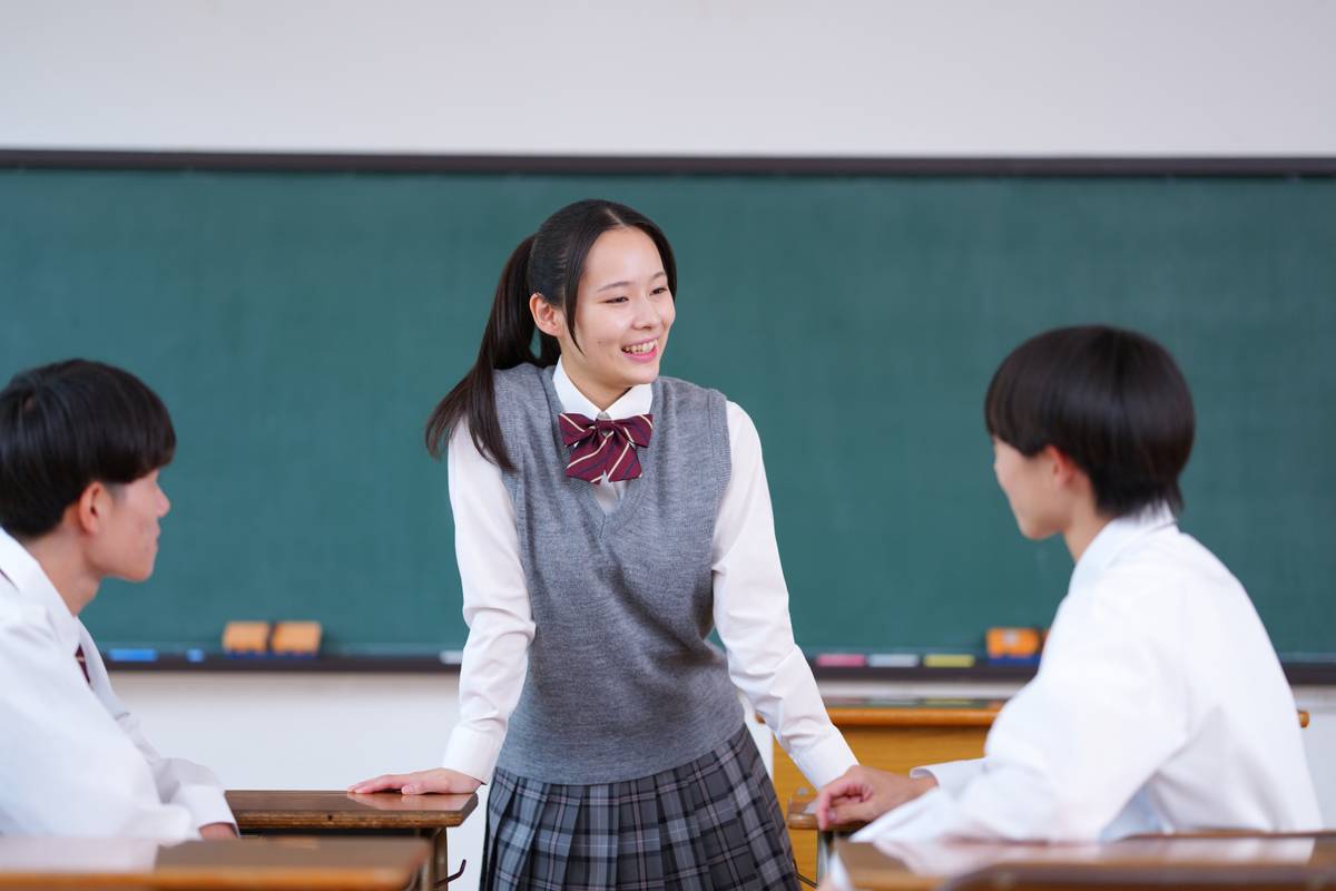 A smiling female student in uniform talks with two male students in a classroom.