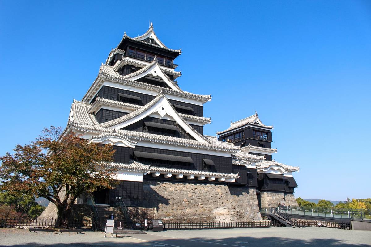 A traditional multi-tiered Japanese castle with black walls and white roofs, on a stone base, under a clear blue sky.