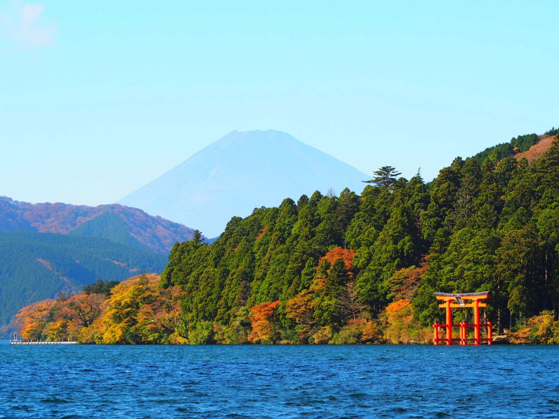 A lake with a red torii gate and autumn trees, with Mount Fuji in the background.