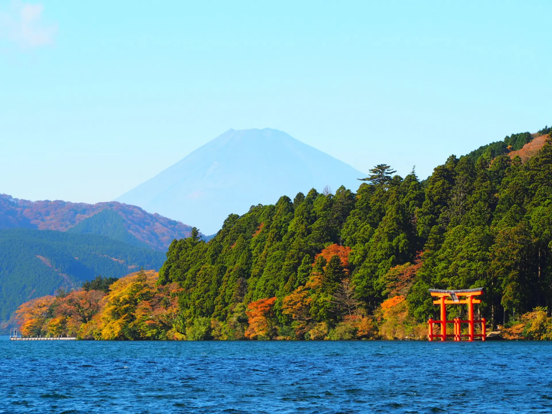 A lake with a red torii gate and autumn trees, with Mount Fuji in the background.