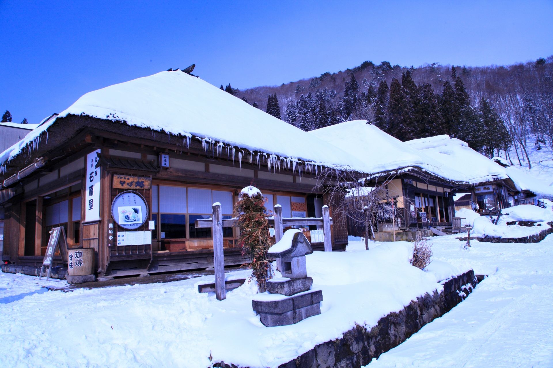 Traditional Japanese buildings with thick snow on thatched roofs and icicles, in a snowy landscape.