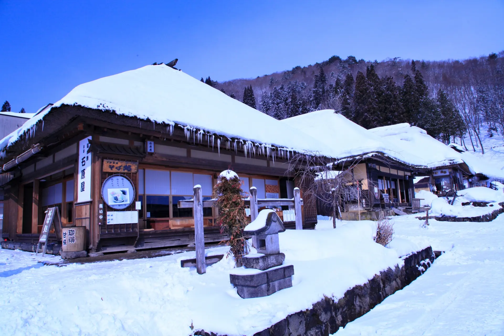 Traditional Japanese buildings with thick snow on thatched roofs and icicles, in a snowy landscape.