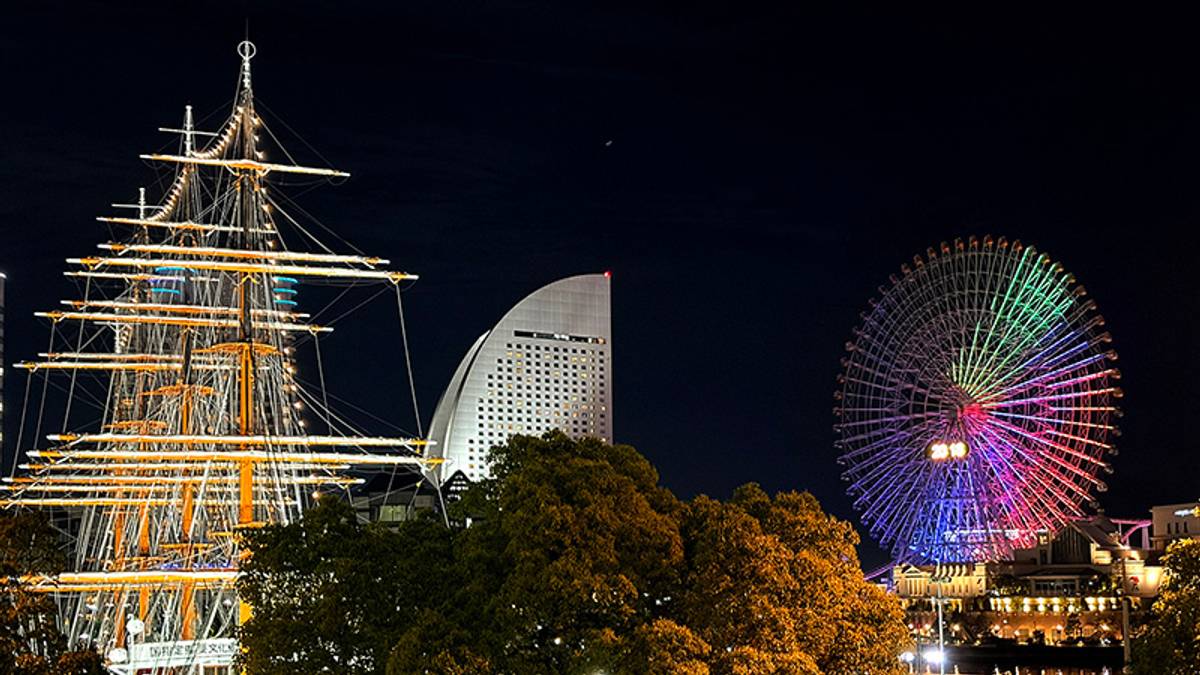 Night scene of an illuminated sailing ship, a curved skyscraper, and a multicolored Ferris wheel.