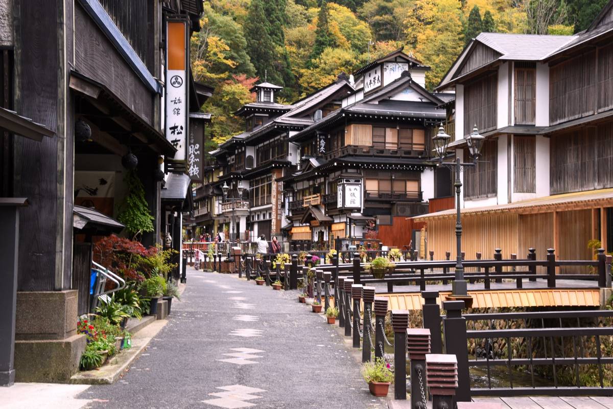 A traditional Japanese street with dark wooden buildings, a river, and colorful autumn trees. 