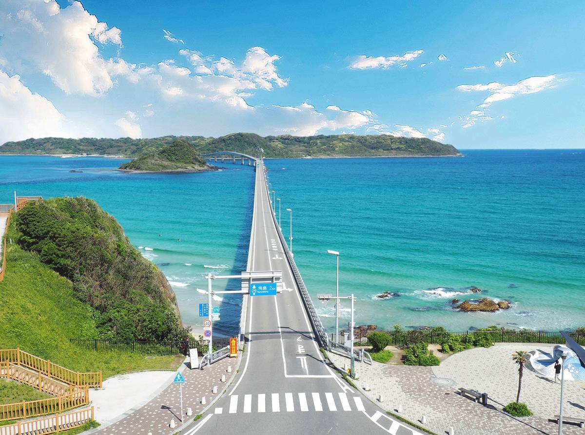 A long bridge extends over clear turquoise ocean towards green islands under a blue sky.