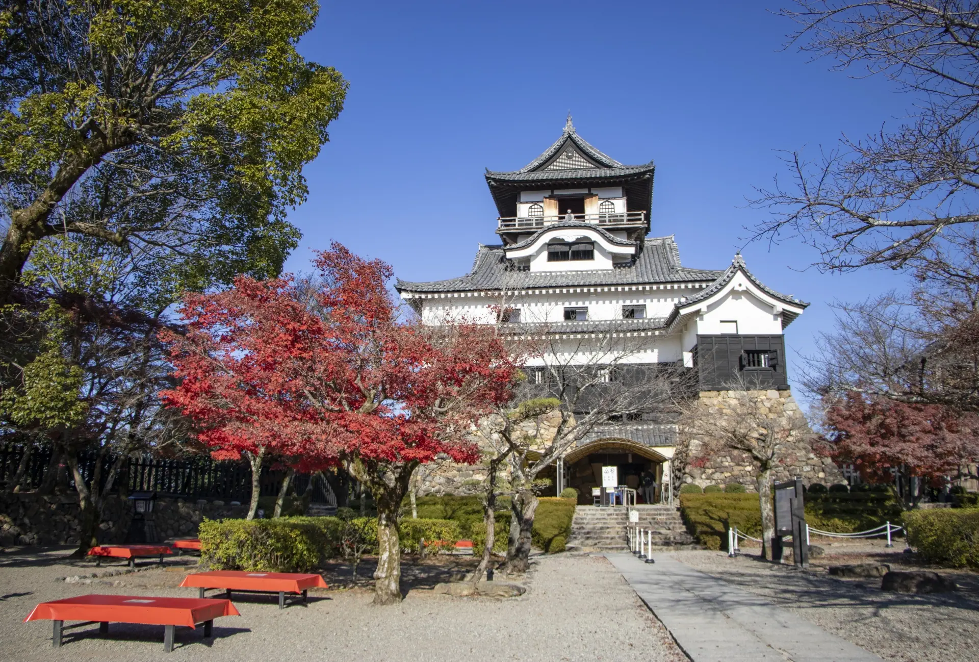 A black and white Japanese castle stands with red autumn leaves, green trees, and red benches under a clear blue sky.