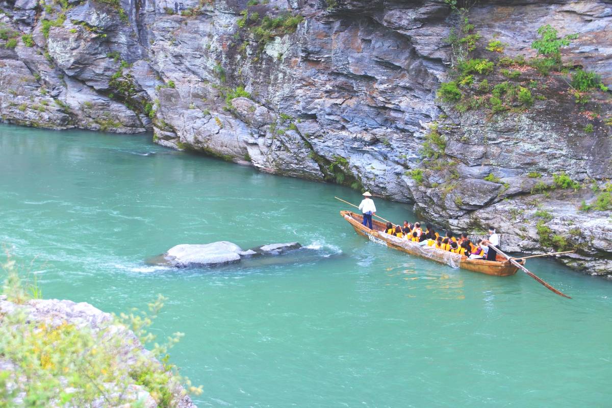 A wooden boat carrying people in yellow life vests navigates a turquoise river between rocky cliffs.