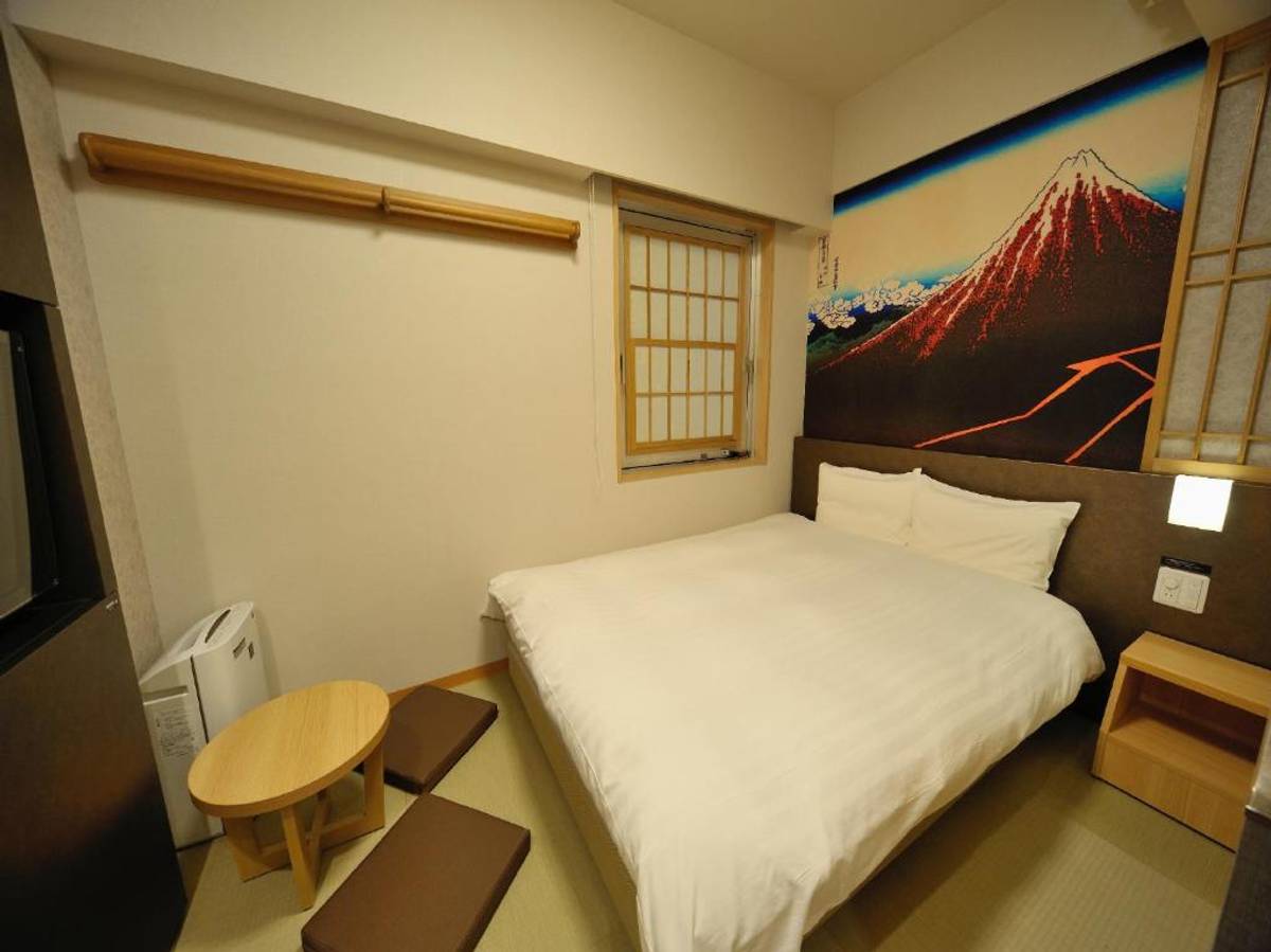 A Japanese-style hotel room featuring a bed, a mural of Mount Fuji above the headboard, a low wooden table with two floor cushions, and a shoji-style window.