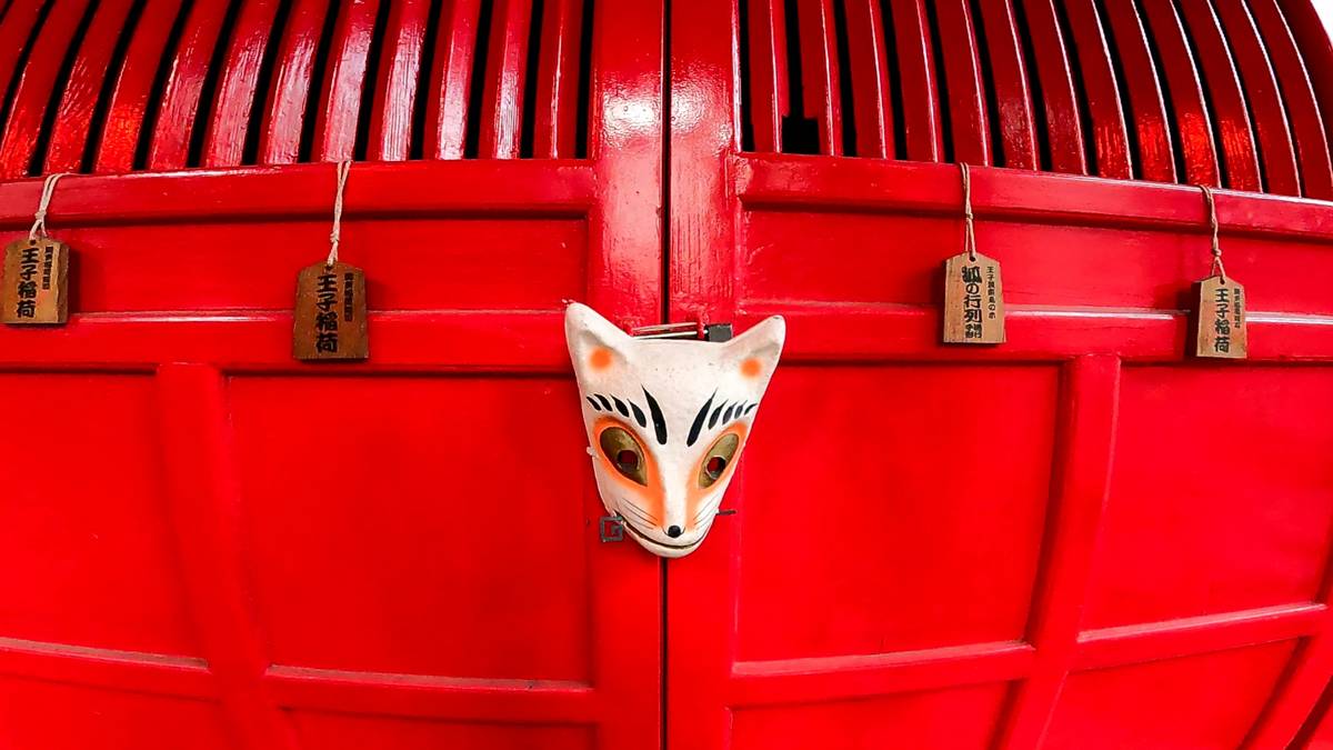 White and orange fox mask on a bright red door with hanging Japanese wooden tags.