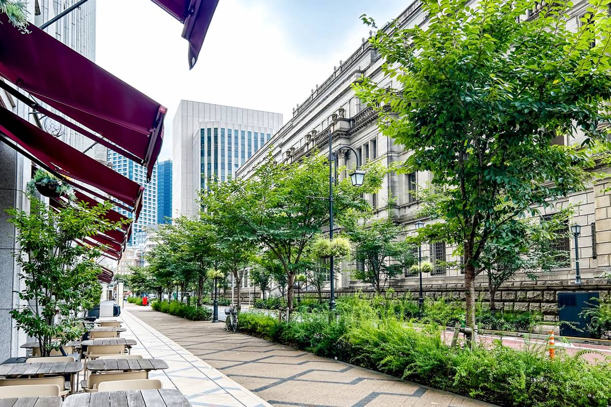 Outdoor cafe tables under red awnings line a city street with trees, a historic stone building, and modern skyscrapers.