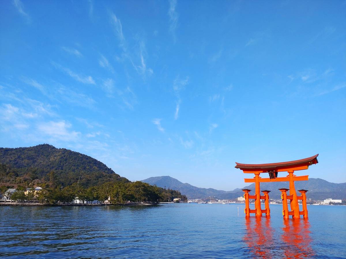 The red-orange Great Torii gate of Itsukushima Shrine rises from the blue water, with forested mountains behind.