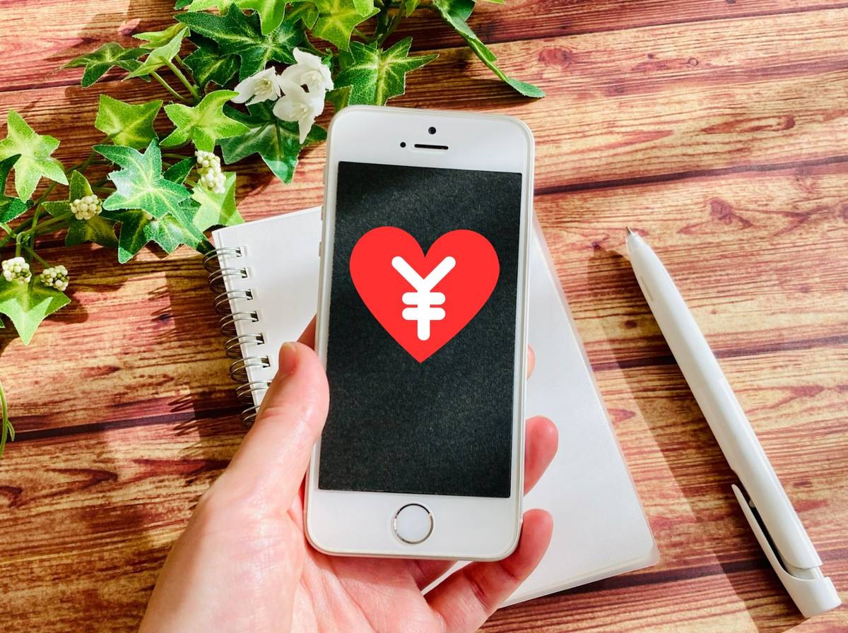 Hand holding a smartphone displaying a red heart with a white Yen symbol, on a wooden table with a notebook, pen, and ivy.