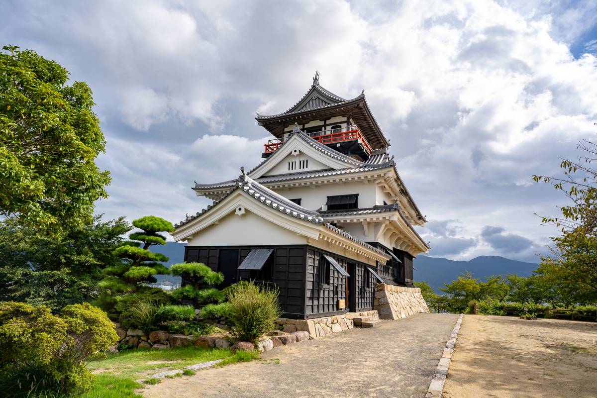 A multi-tiered Japanese castle with white walls and dark roofs stands amidst green trees under a cloudy sky.