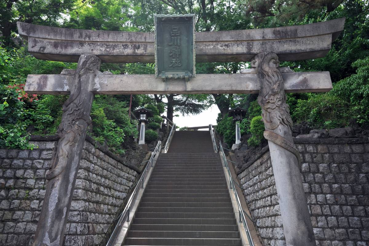 Stone torii gate at the bottom of a long staircase, flanked by stone walls and green trees.