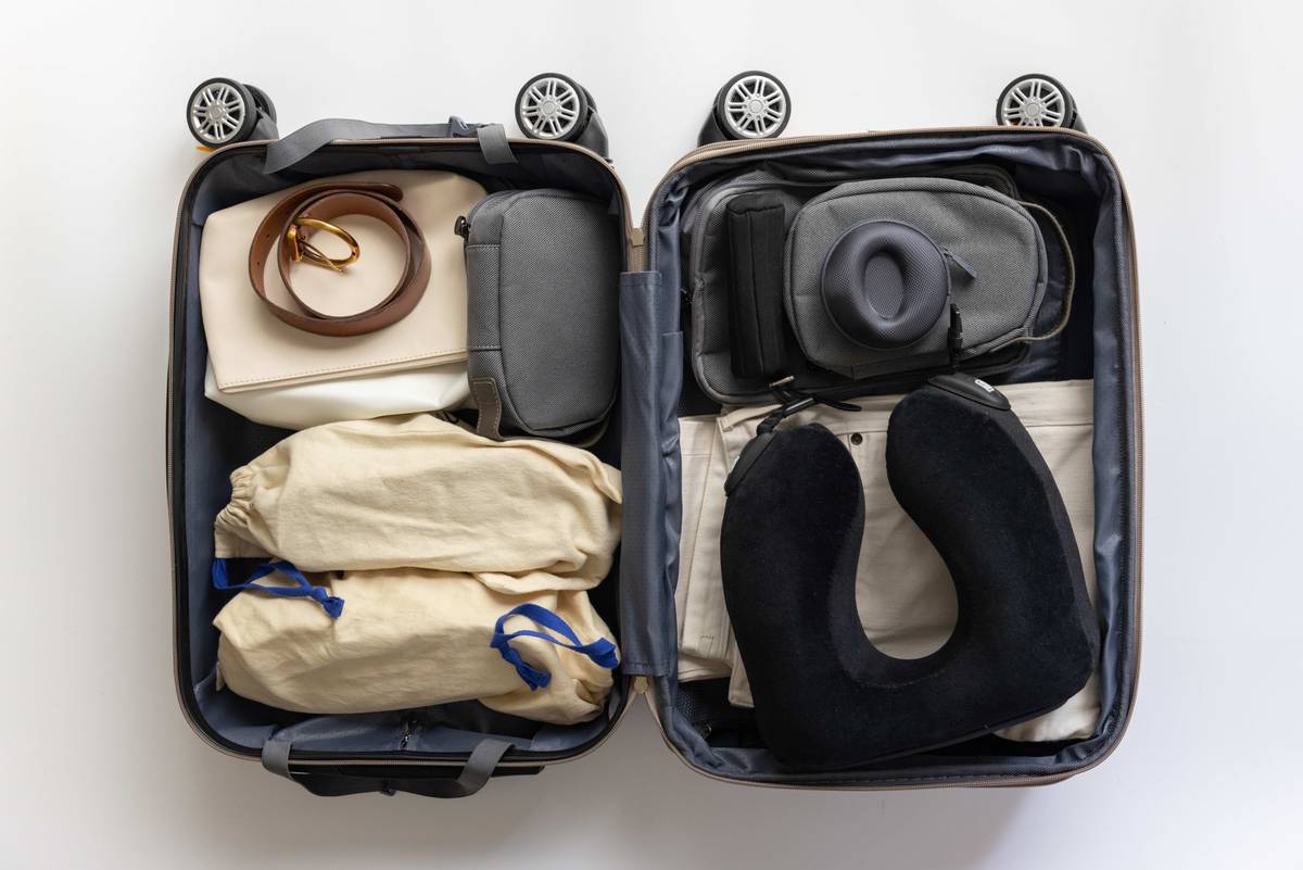 An open suitcase neatly packed with clothing bags, a belt, a tech pouch with headphones, and a neck pillow.