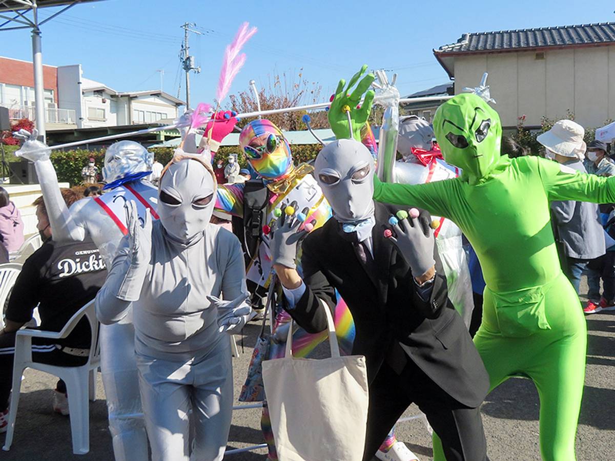 A group of people in diverse alien costumes, including green, gray, and silver, posing at an outdoor event.