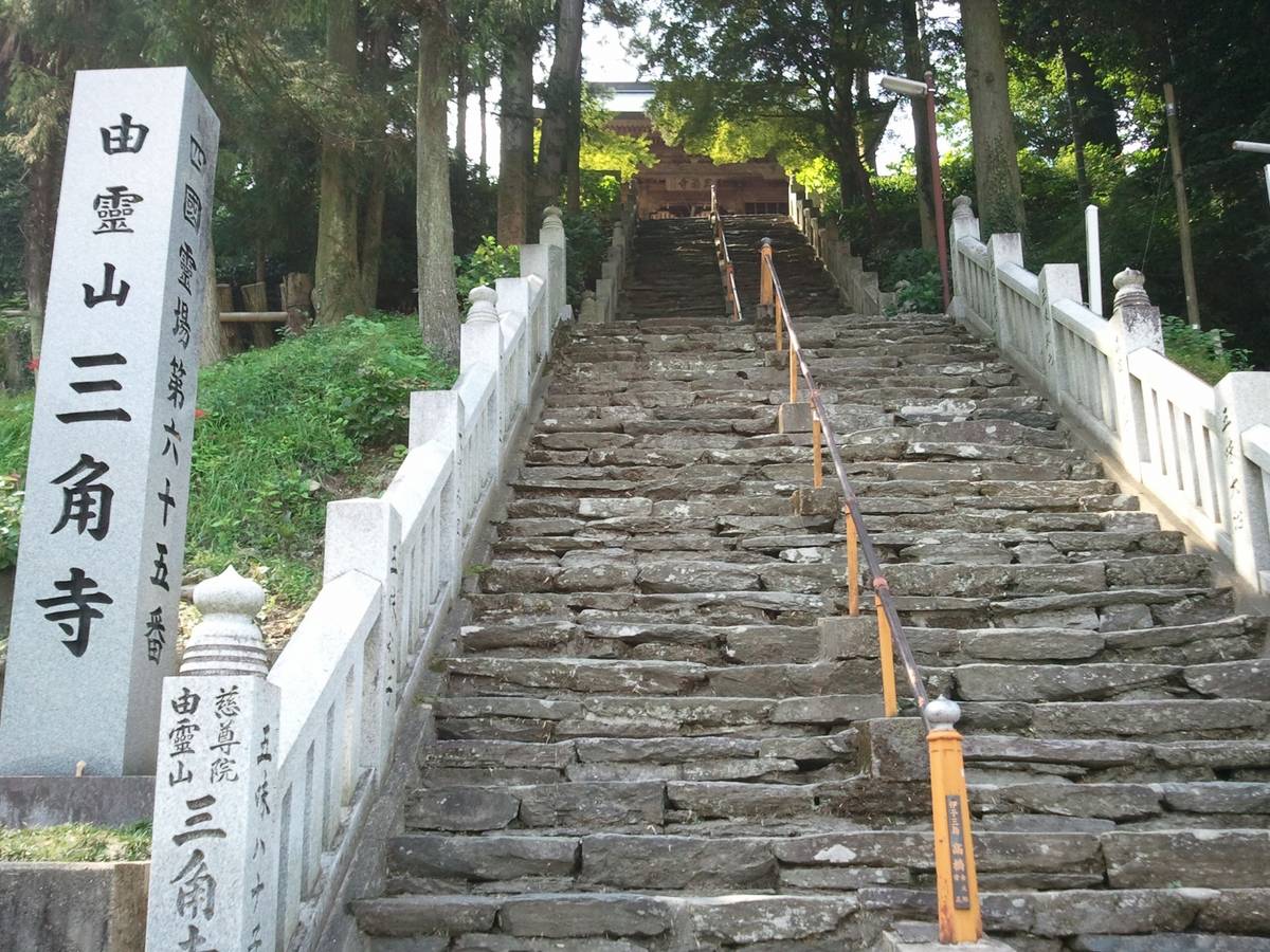 A steep stone staircase with white railings, an orange handrail, and a monument with Japanese text, leading up to a building through dense trees.