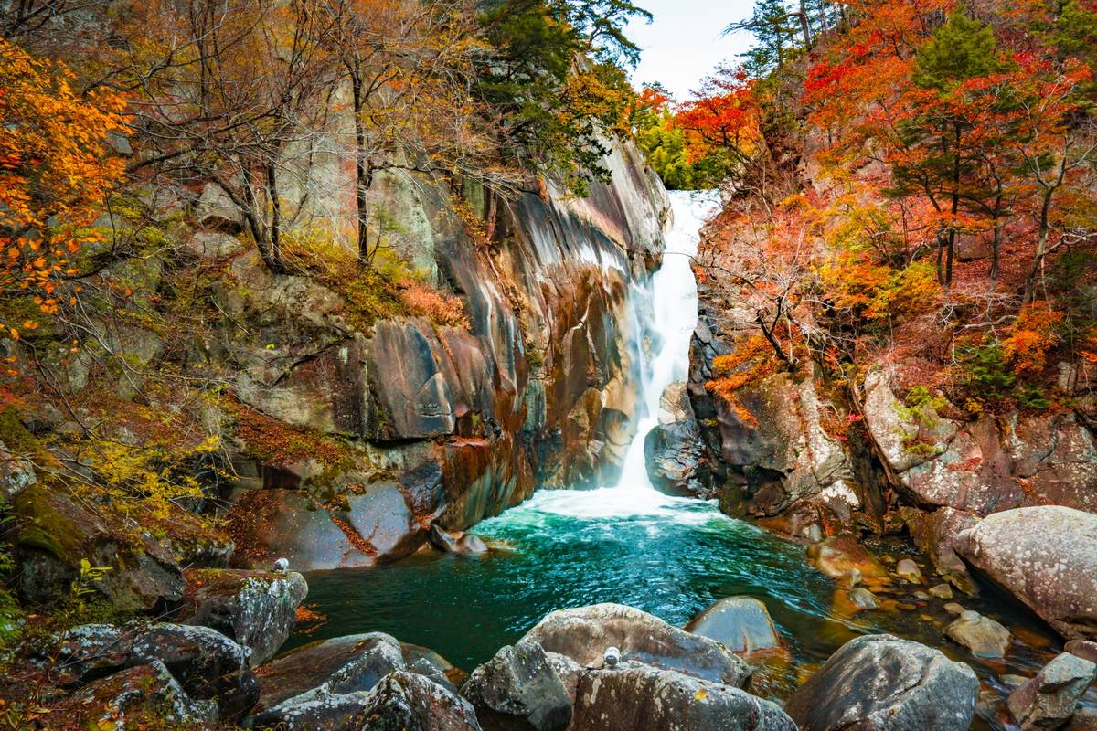 Powerful waterfall plunges into a vibrant turquoise pool, surrounded by colorful autumn foliage and rocky cliffs.