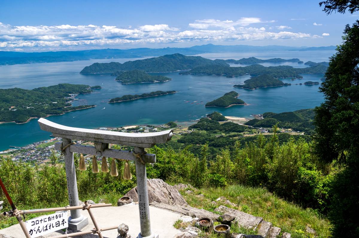 A stone torii gate with bells overlooks a scenic bay dotted with many green islands under a blue sky.