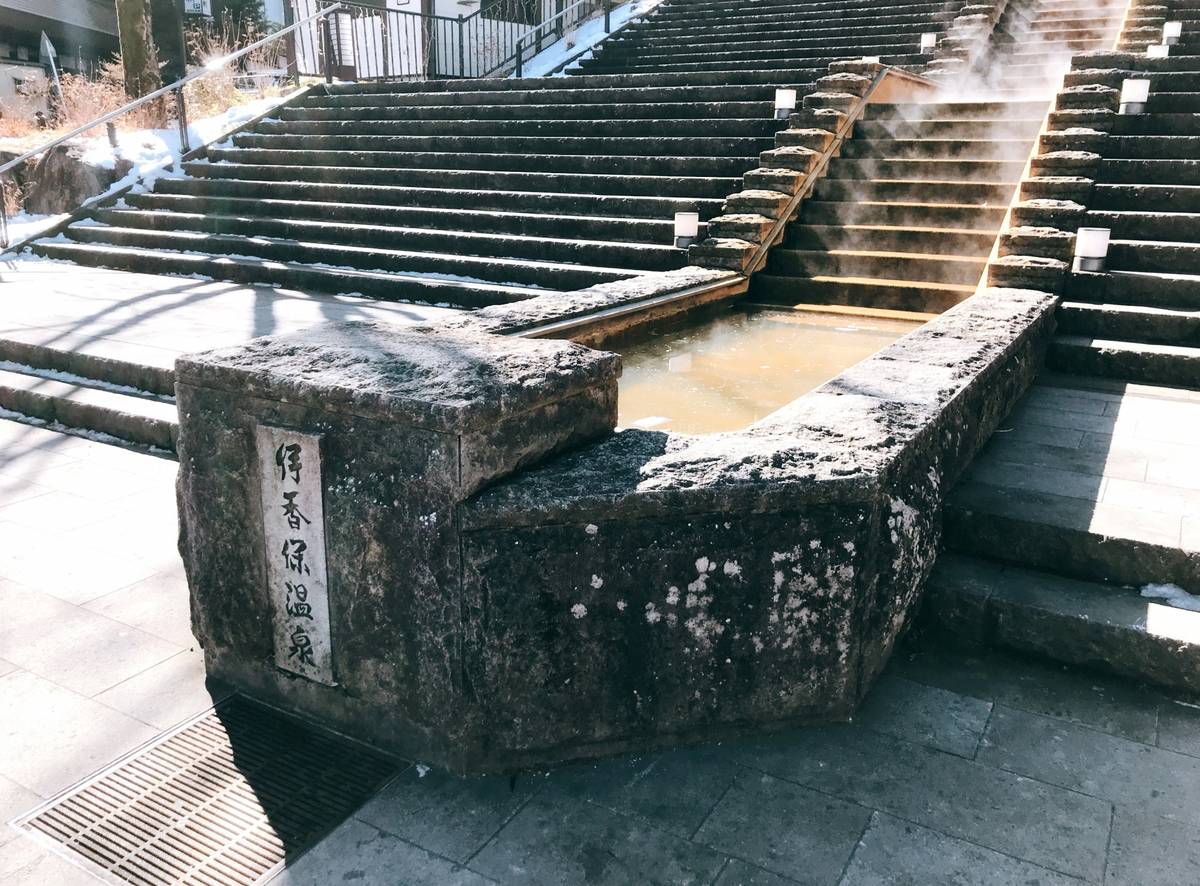 Steaming hot spring water in a stone channel and pool beside a large, snow-dusted stone staircase.