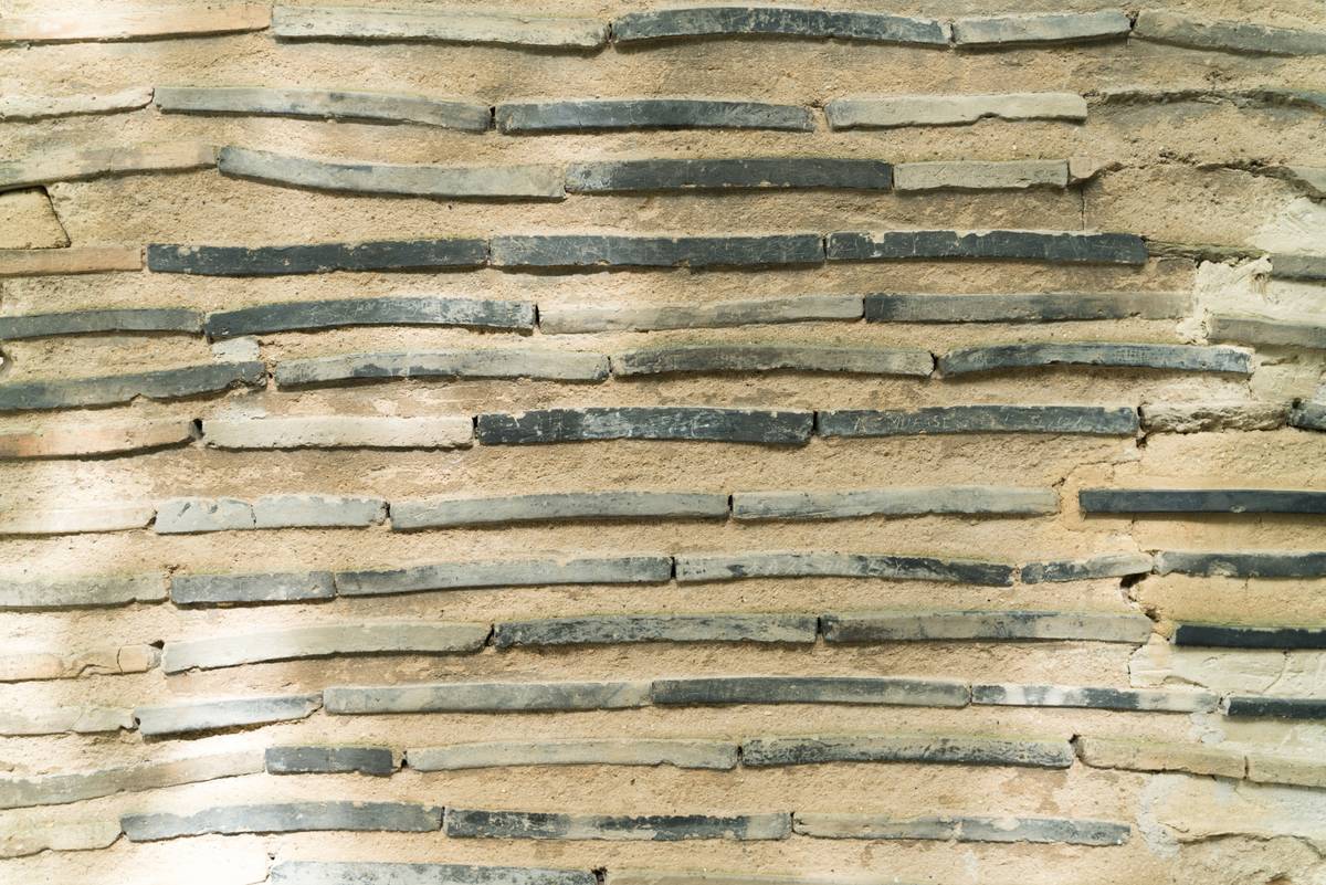 Wall texture made of irregular horizontal dark grey tile fragments set in light brown mortar.