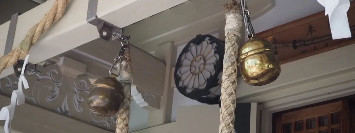 Brass bells and thick ropes hang from a shrine's wooden eaves, with white paper strips and a circular emblem.