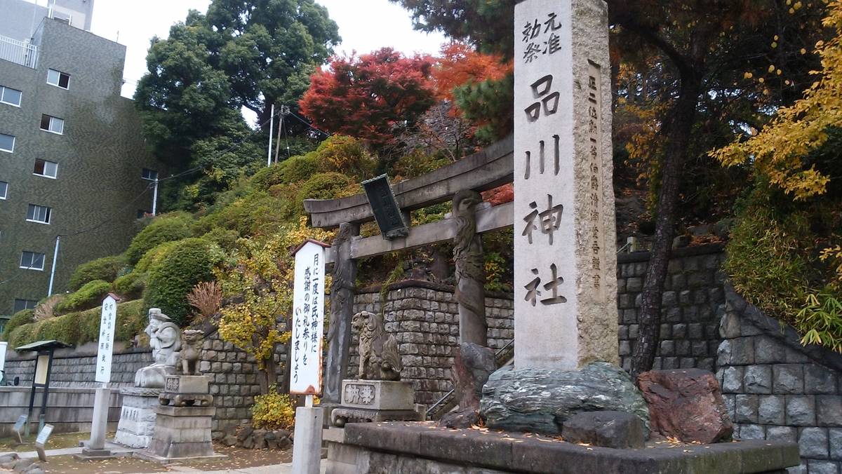 Entrance to Shinagawa Shrine with a large stone pillar, guardian statues, and colorful autumn foliage on a hillside.