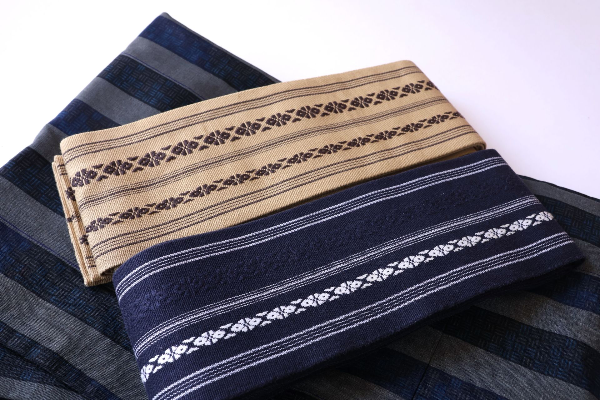 Two folded patterned textile sashes, one beige and one dark blue, on a striped blue fabric.
