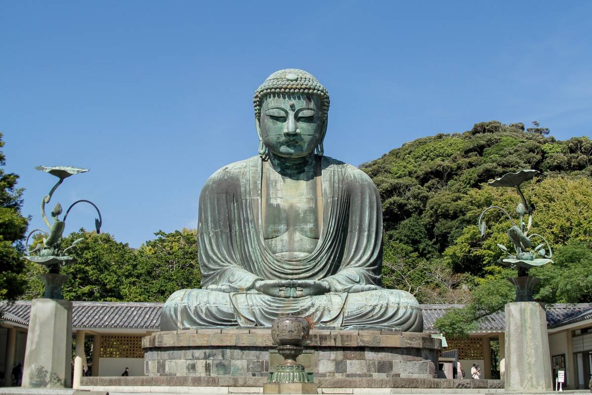 A large, weathered bronze statue of Buddha seated outdoors, surrounded by green trees under a clear blue sky.