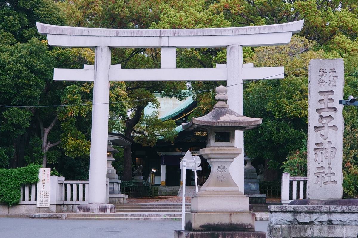 White torii gate marking the entrance to a Japanese shrine, with a stone lantern and carved monument in the foreground.