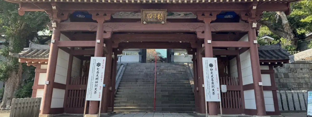 A traditional Japanese temple gate made of reddish-brown wood, with stone steps leading through it and up.