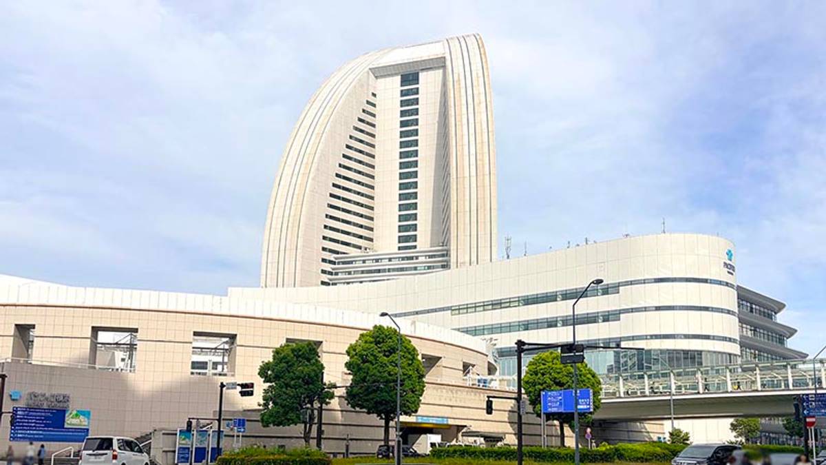 A tall, sail-shaped skyscraper overlooks modern buildings, a street, and a pedestrian bridge.