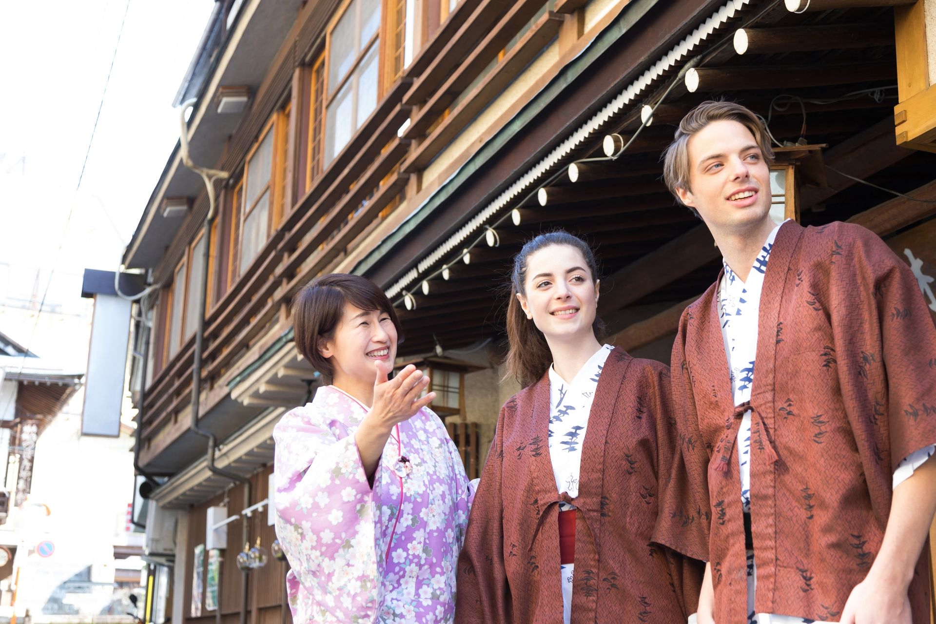 A Japanese woman in a pink kimono smiles and gestures next to two young people in brown yukatas on a traditional street.