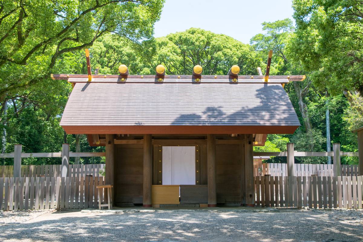 Traditional wooden Japanese shrine with a gray roof, golden roof ornaments, and lush green trees.