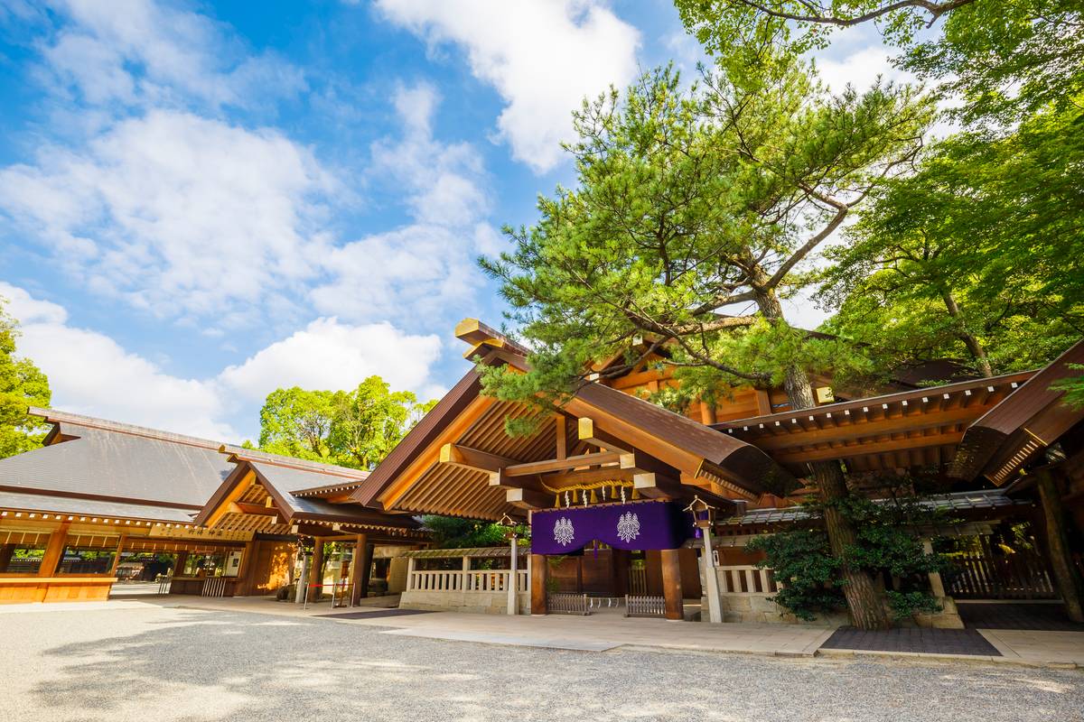 Traditional Japanese shrine complex with wooden architecture, a purple banner, and lush trees under a blue sky.