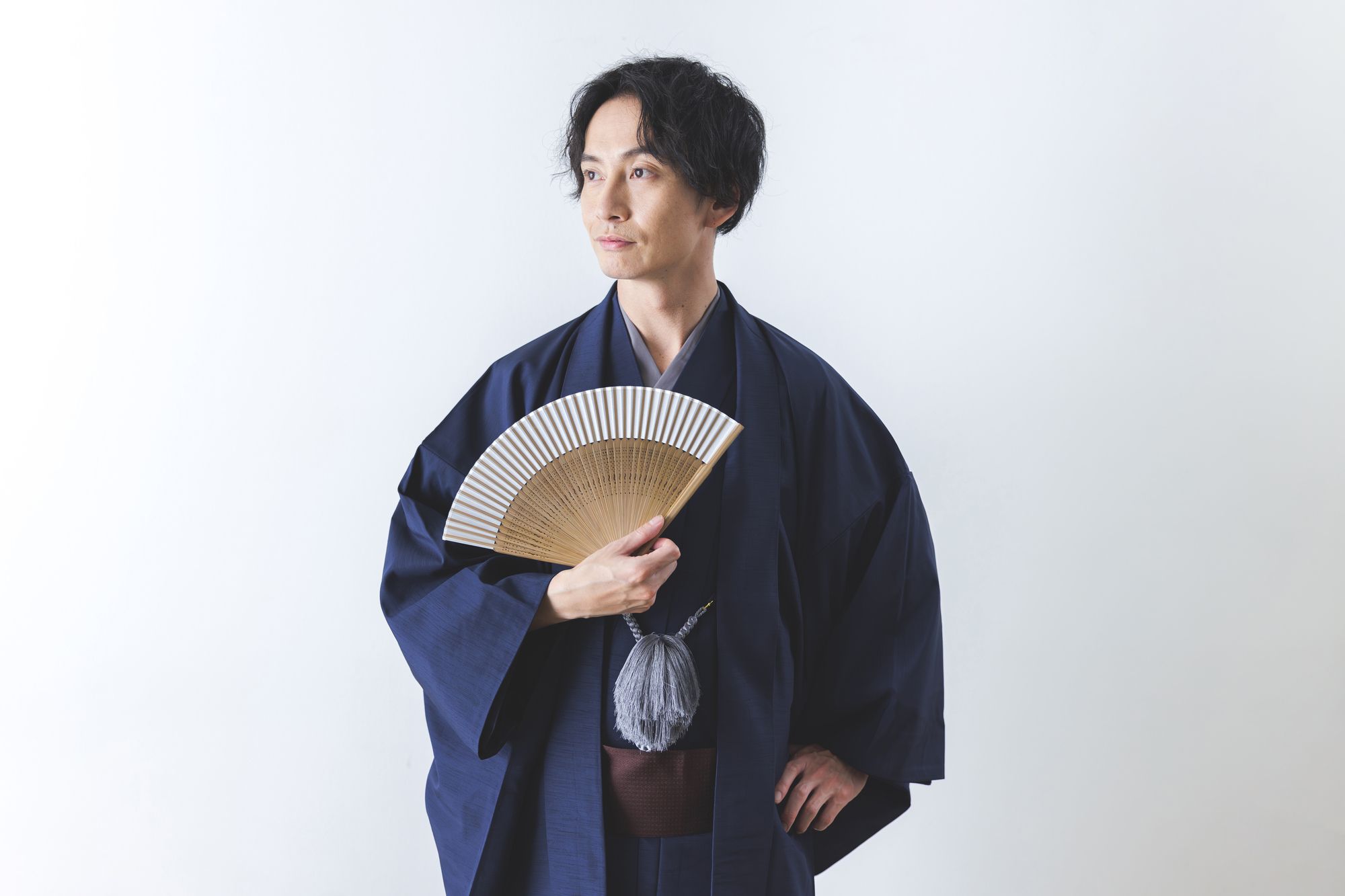 A man in a dark blue kimono holds a folding fan and looks to the side against a white background.