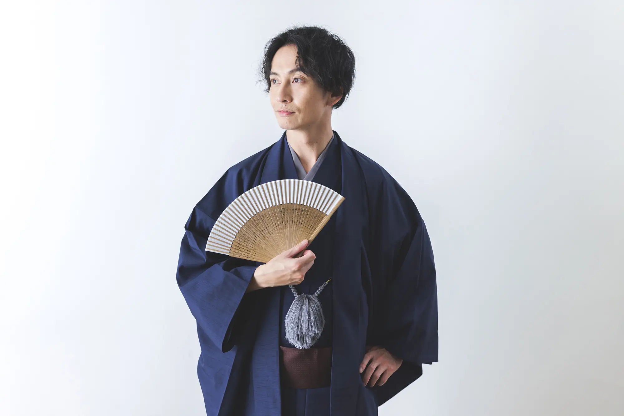 A man in a dark blue kimono holds a folding fan and looks to the side against a white background.