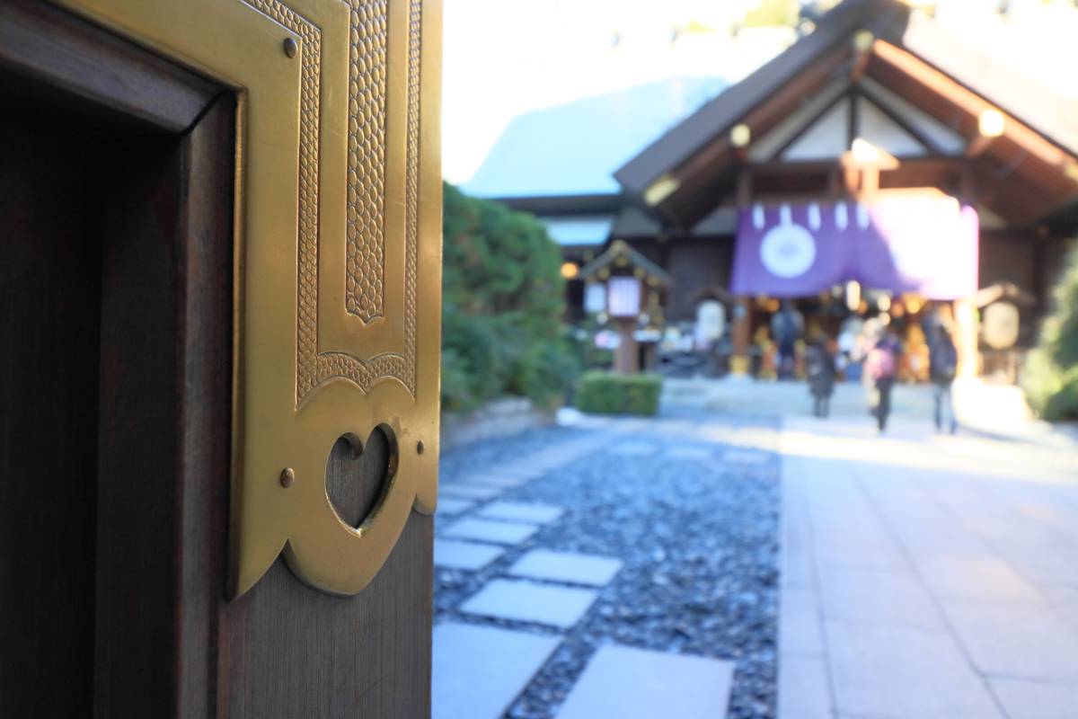 Ornate brass door decoration with a heart cutout, overlooking a blurred Japanese shrine and people.