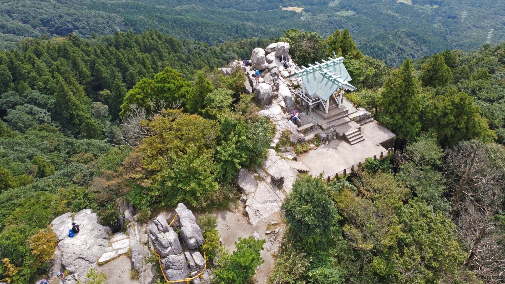 Aerial view of a rocky mountain peak with a modern pavilion, people, and dense forest.
