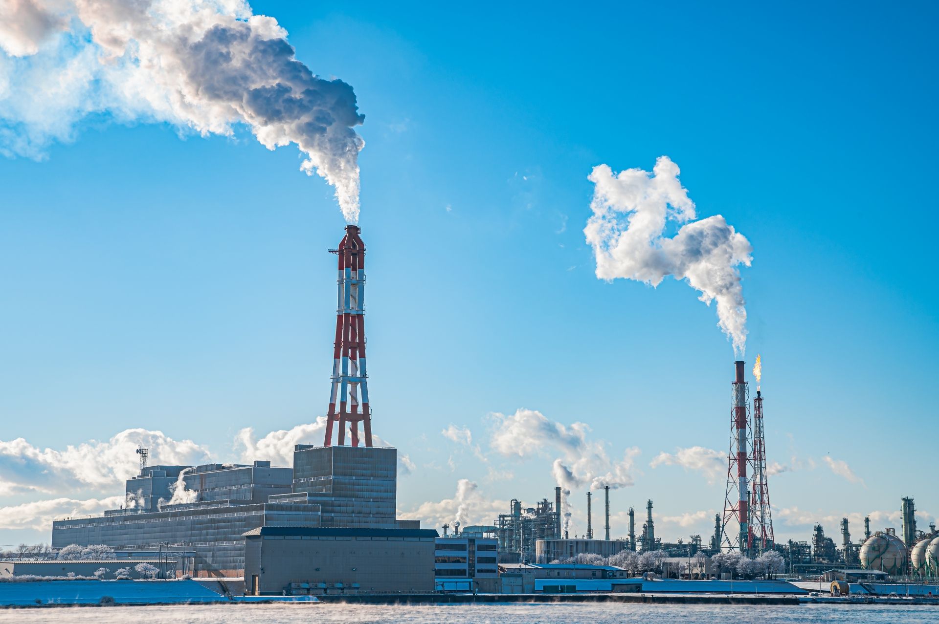 Industrial plant with tall smokestacks emitting plumes of white smoke into a clear blue sky.