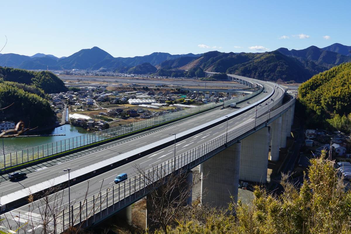 A long, elevated highway bridge with cars curves through a valley with a town, river, and mountains.